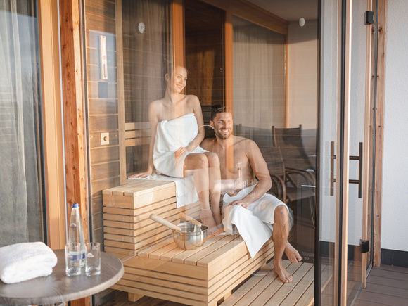 Paar sitzt lachend im Bademantel in moderner Holzsauna, umrandet von Glaskasten