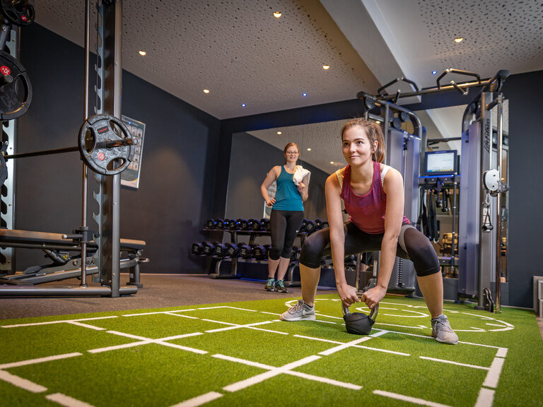 Frau bei Hebeübung mit Kugelgewicht auf grünem Teppich in Fitnessraum mit Spiegelwand