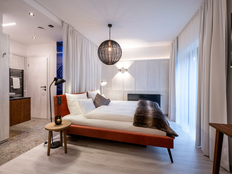 Doppelbett in modernem, hellen Zimmer, darüber runde Lampe, dahinter offenes Badezimmer