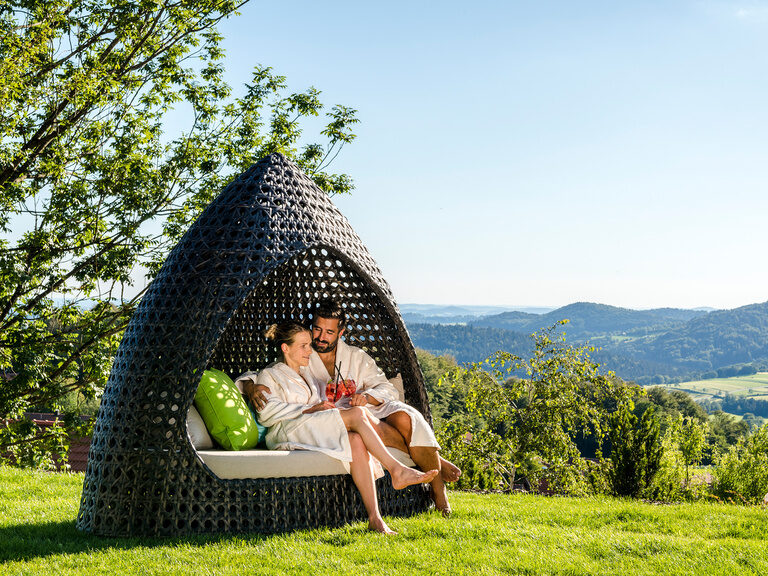 Paar in Bademantel mit Getränken in Lounge-Insel in sonnigen Garten mit Landschaftsblick