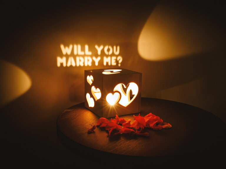 Teelicht in Herz-Holzbox, Rosenblätter und 'Will you marry me?'-Lichtschrift an der Wand