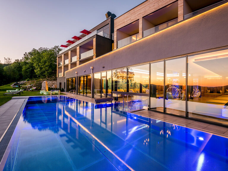 Außenansicht vom 4-Sterne-Wellnesshotel Hüttenhof mit Außenpool bei Sonnenschein Außenansicht Hotel mit Außenpool, Liegen und Sonnenschirme auf Wiese mit Wald im Hintergrund