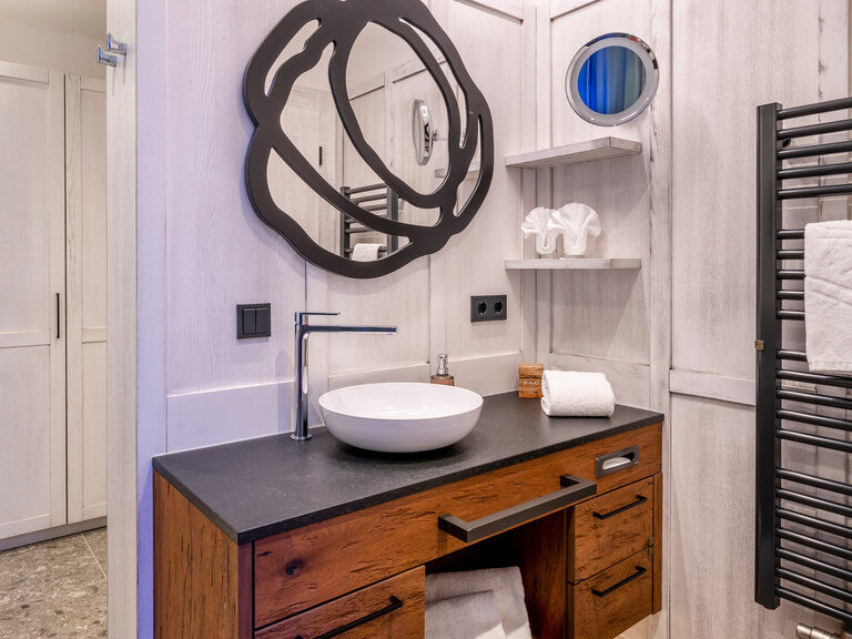 Bad mit modernem Waschbecken auf Holzwaschtisch und dunkler Heizung, darüber abstrakter Spiegel