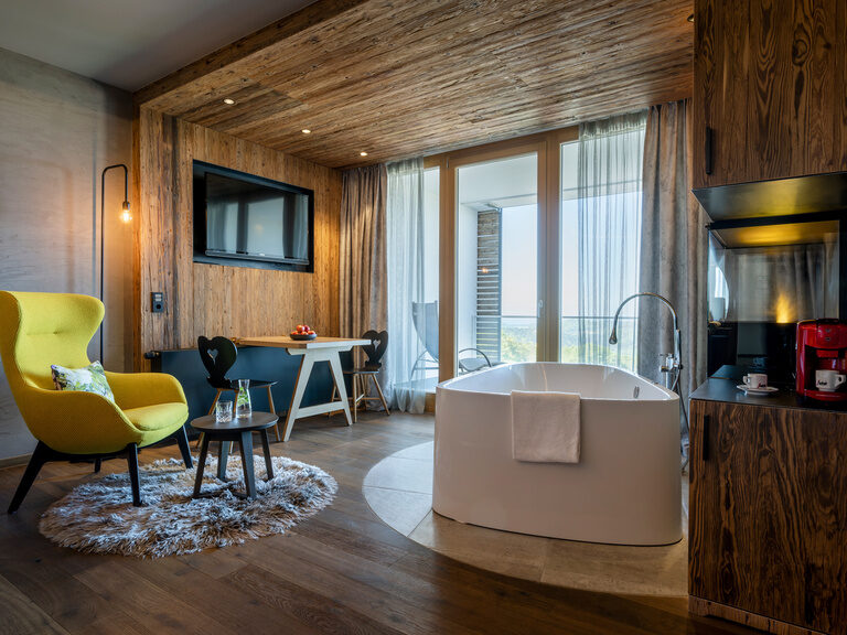 Stilvolle Suite mit Sitzbereich, TV an Holzwand, Balkon mit Liege und frei stehender Badewanne