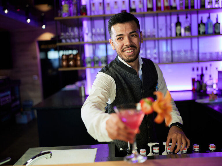 Barkeeper in violett leuchtender Bar überreicht bunten Cocktail im Hüttenhof