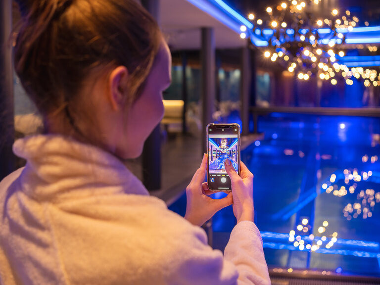 Frau fotografiert mit Handy den beleuchteten Innenpool im modernen 4-Sterne-Hotel Hüttenhof