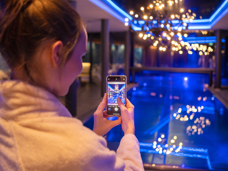 Frau fotografiert mit Handy den beleuchteten Innenpool im modernen 4-Sterne-Hotel Hüttenhof