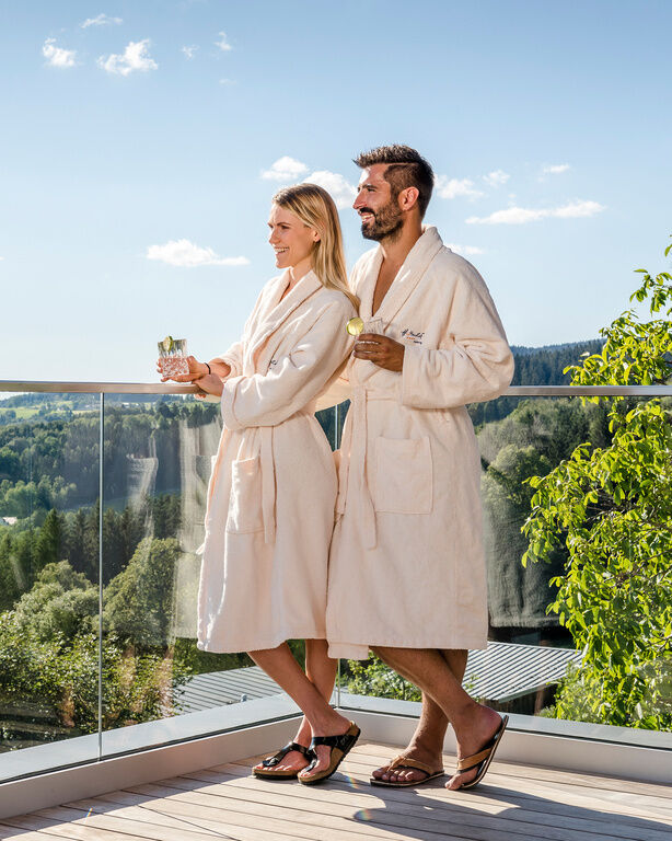 Lachendes Paar steht im Bademantel und Getränken in der Hand auf Terrasse vom Hüttenhof