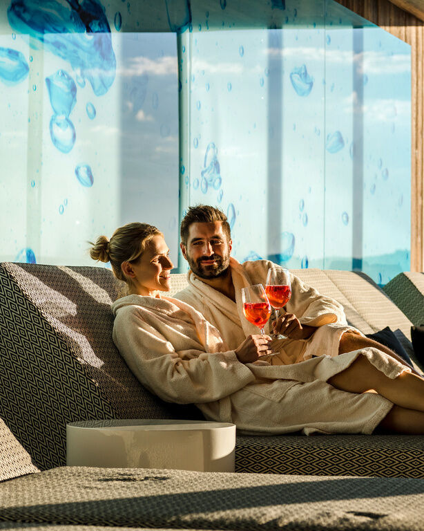Paar im Bademantel liegt lachend mit Cocktails auf Sofa-Liege in Panorama-Ruheraum