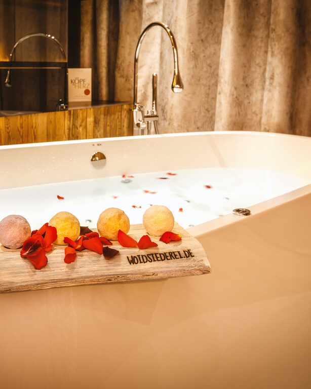 Befüllte Badewanne vor Kamin, mit Rosenblättern, darauf Holzbrett mit runden Badebomben