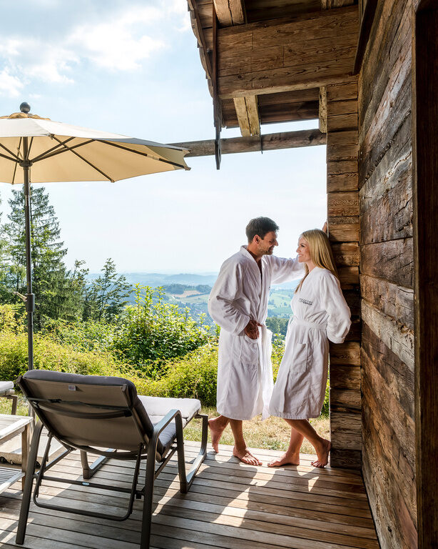 Pärchen steht auf der Terrasse des Chalets mit Whirlpool und gemütlichen Liegen, traumhafter Blick auf den bayerischen Wald