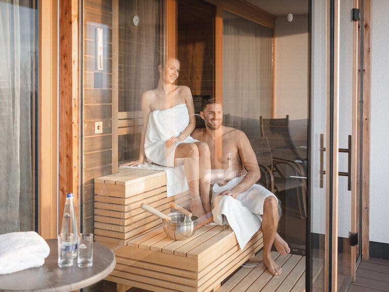 Lachendes Paar sitzt in der Sauna des Hotel Hüttenhof und genießt den Ausblick durch die Glastür