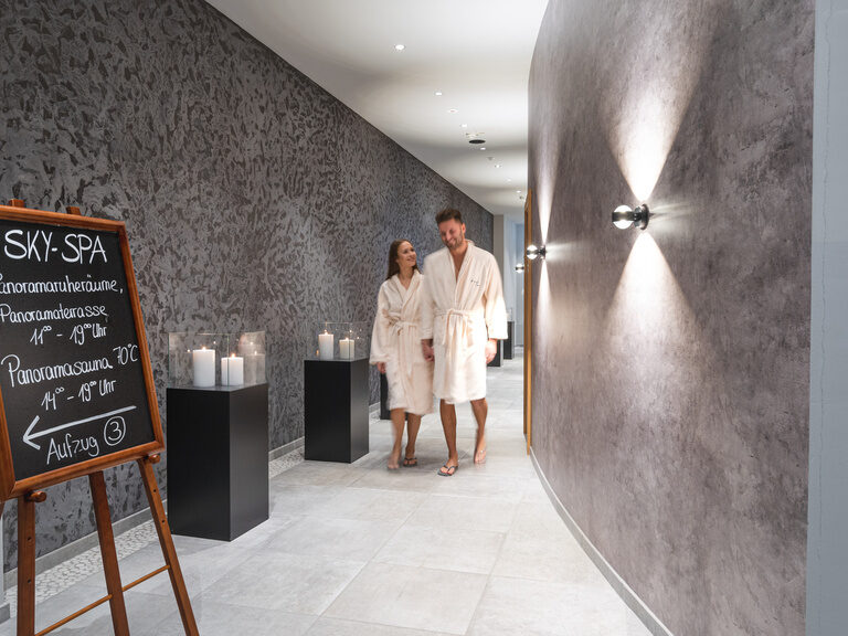 Paar im Bademantel geht Gang Richtung Sky Spa im Luxushotel Hüttenhof Paar im Bademantel schlendert Gang entlang, ein Schild weist den Weg zum Sky-Spa
