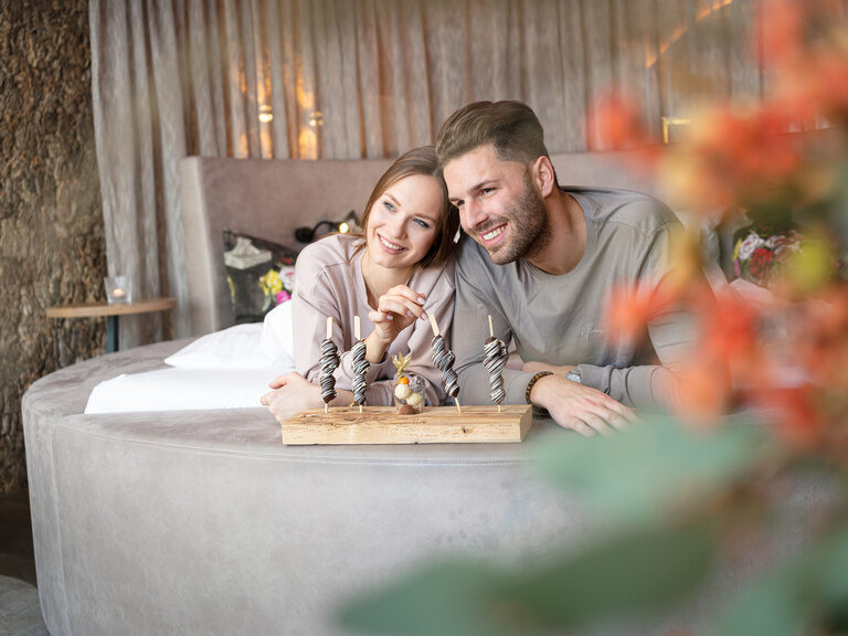 Paar lachend mit Schoko-Frucht-Spießen im Luxushotel Hüttenhof Paar liegt lachend auf Bett mit Schoko-Frucht-Spießen im Luxushotel Hüttenhof