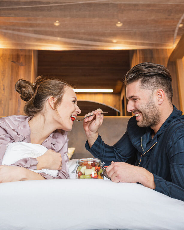 Mann reicht Frau lächelnd einen Löffel mit Obst beim Frühstück im Bett im Hüttenhof