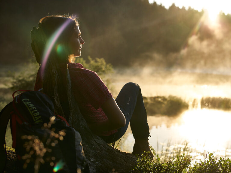 Frau mit Rucksagt sitzt vor sonnigem Sumpf im Bayerischen Wald