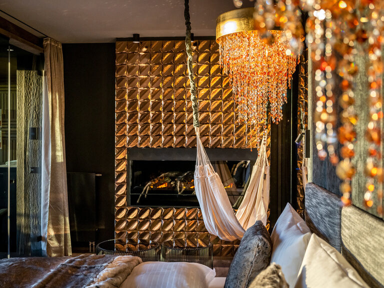 Goldener Kamin und Hängematte in moderner Luxussuite im Wellnesshotel Hüttenhof Goldener, glänzender Kamin, Hängematte, Kronleuchter und modernes Doppelbett in Luxussuite