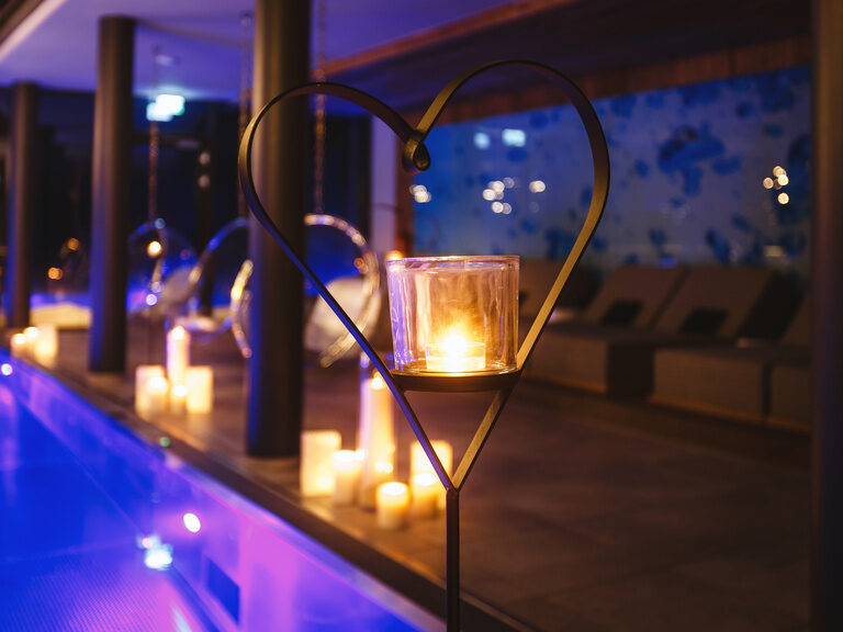 Windlicht in herzförmiger Stahlhalterung, im Hintergrund beleuchteter Innenpool umgeben von Kerzen