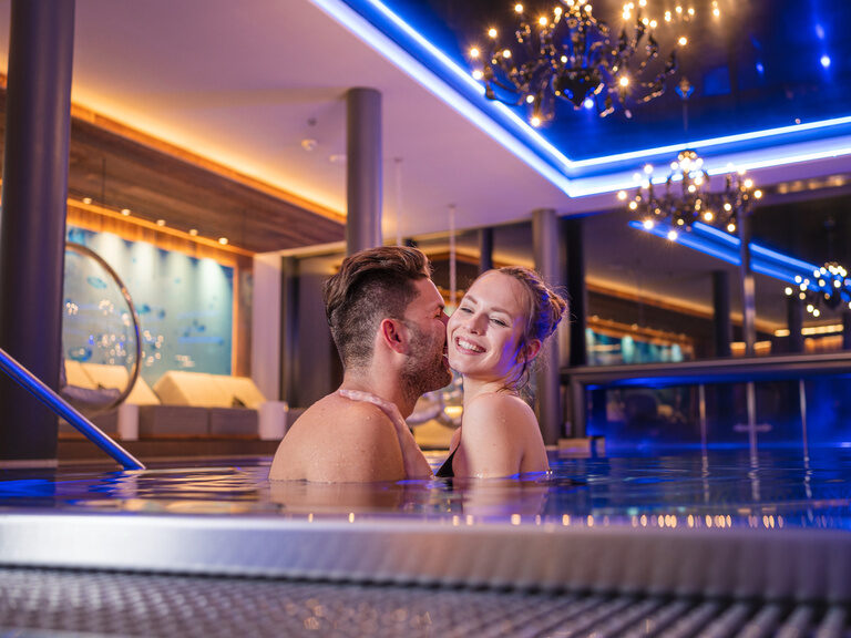 Küssendes Paar sitzt im Schlafanzug mit einem Glas Sekt im gemütlichen Bett des 4 Sterne Wellnesshotel Hüttenhof im Bayerischen Wald
