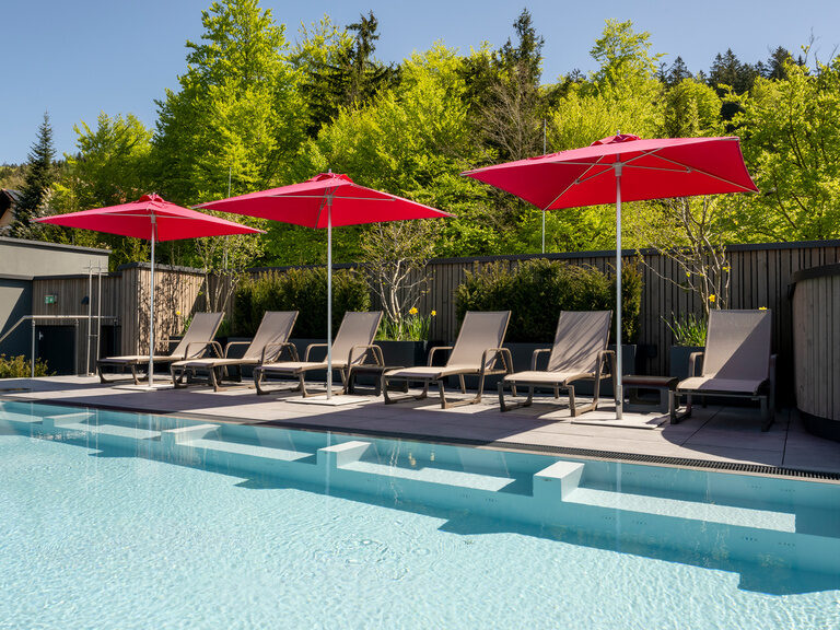 Liegen mit Sonnenschirmen neben Dachpool vom 4-Sterne-Luxushotel Hüttenhof. Liegen und farbenfrohe Sonnenschirme neben dem Dachpool, im Hintergrund der grüne Wald