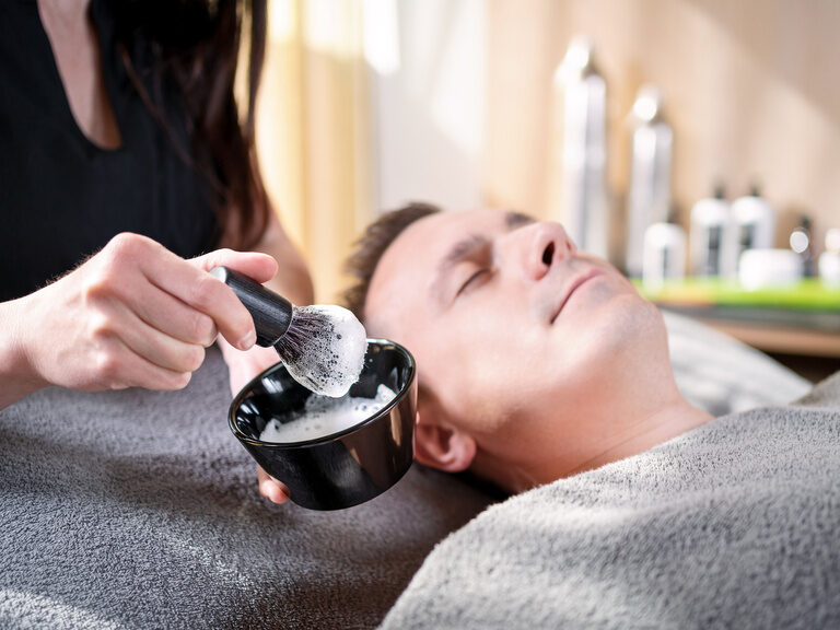 Pinsel wird in Gesichtsmaske getaucht, im Hintergrund lächelnder Mann auf Behandlungsliege