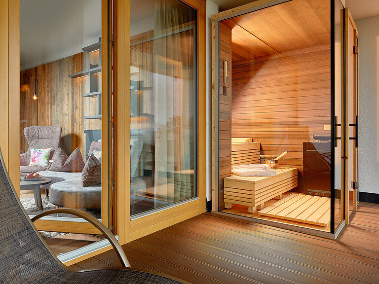 Außenansicht von beleuchteter, modernen Holz-Sauna umrandet von Glaskasten mit dunklen Türgriffen