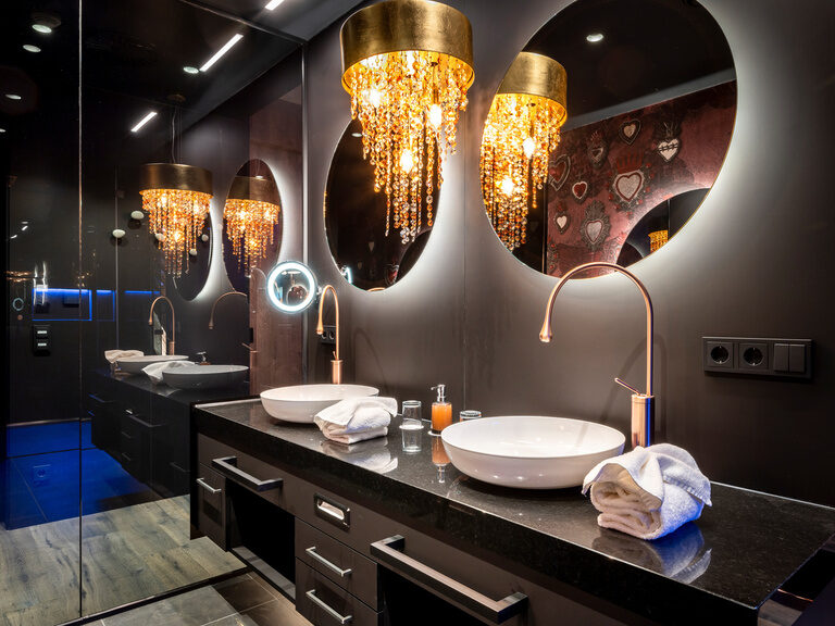Modernes, dunkles Bad mit runden Spiegel und Kronleuchter im Wellnesshotel Hüttenhof Modernes, dunkles Bad mit runden Spiegeln und Waschbecken, leuchtendem Kosmetikspiegel und Kronleuchter