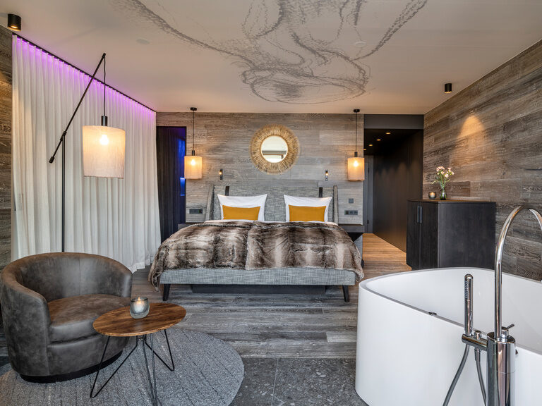 Modernes Hotelzimmer mit Doppelbett, Badewanne und Sitzbereich im Luxushotel Hüttenhof Modernes Hotelzimmer mit Holzwänden, Doppelbett, frei stehender Badewanne und Ledersessel