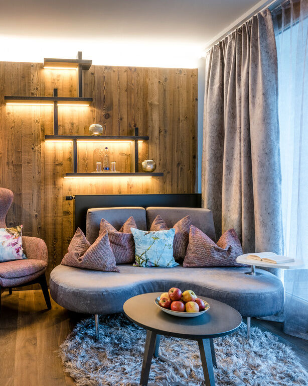 Rundes Sofa mit dunklem, rundem Sofatisch, daneben Stoff-Sessel, an Holzwand mit beleuchtetem Regal