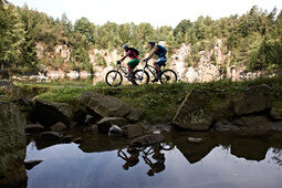 Zwei Radfahrer auf Mountainbikes fahren am Ufer eines Sees im Bayerischen Wald entlang, mit Felsen im Hintergrund.