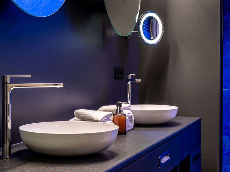 Zwei Runde Waschbecken auf dunklem Waschtisch, darüber leuchtender Kosmetikspiegel in modernem Badezimmer