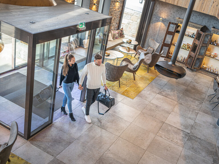 Paar betritt durch automatische Glastür den Eingangsbereich mit Couch im Wellnesshotel Hüttenhof