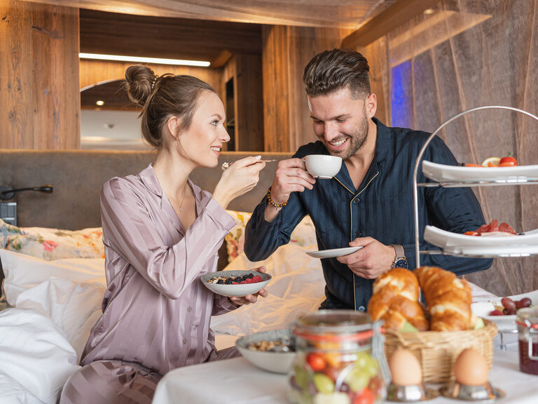 Paar genießt Frühstück im Bett im 4-Sterne-Wellnesshotel Hüttenhof Paar genießt lächelnd Frühstück im Bett mit Croissants, Kaffee und Obst
