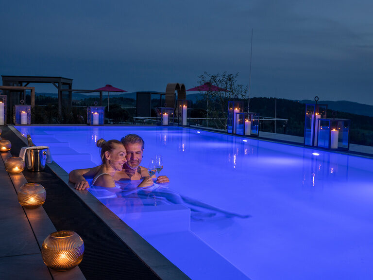 Paar in beleuchteten Dachpool nach Sonnenuntergang im Luxushotel Hüttenhof Paar stößt mit zwei Weingläsern in beleuchteten Dachpool nach Sonnenuntergang an