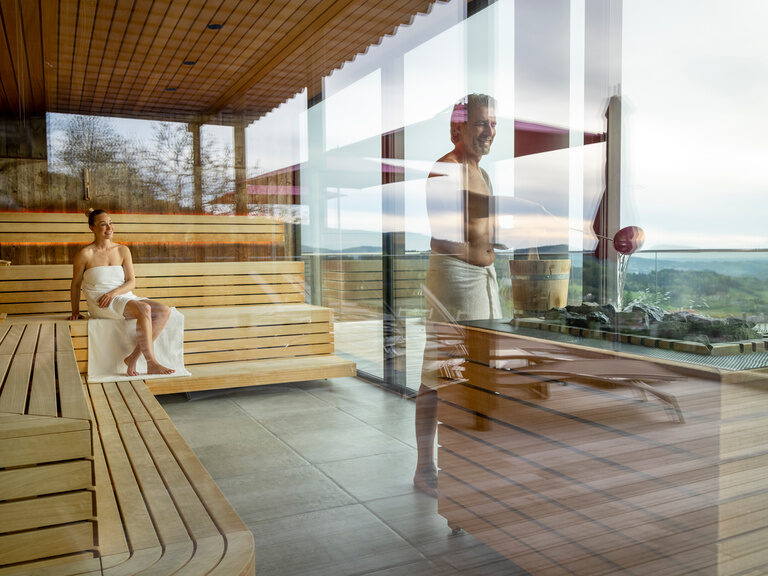 Paar in Panoramasauna des 4-Sterne-Wellnesshotel Hüttenhof Paar genießt die moderne Panoramasauna, sichtbar hinter der Glastür