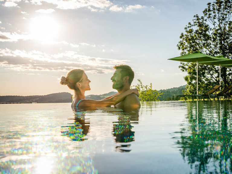 Paar ist lachend und Arm in Arm im Außenpool vom Hotel Hüttenhof Paar lacht Arm in Arm im Außenpool neben Wiese mit Liegen bei Sonne