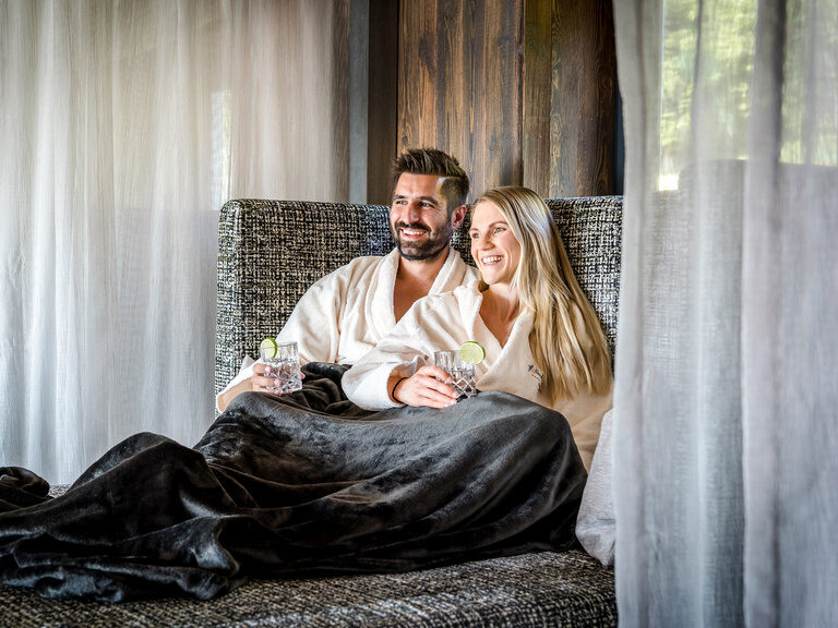 Paar liegt lachend auf einer Sofa-Liege und genießt Erfrischungsgetränke im Hotel Hüttenhof