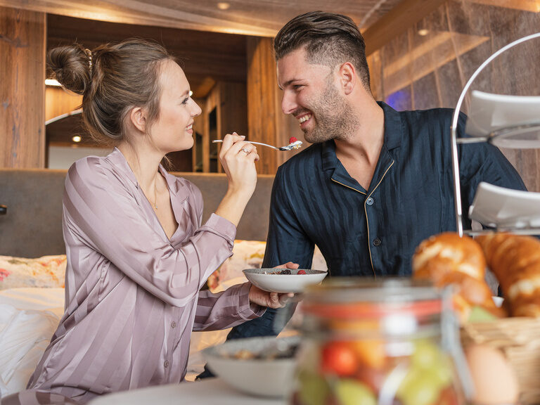 Lachendes Paar genießt ein gemütliches Frühstück im Bett im Luxushotel Hüttenhof Fröhliches Paar genießt das Frühstück im Bett und Frau hält dem Mann liebevoll einen Löffel hin