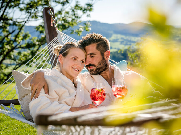 Paar liegt Arm in Arm in Hängematte im Grünen im 4-Sterne-Hotel Hüttenhof Paar liegt lächelnd Arm in Arm in Hängematte im Grünen mit Cocktails in der Hand