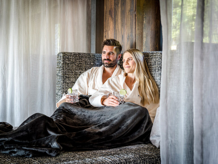 Paar im Bademantel liegt auf einer Sofa-Liege mit Erfrischungsgetränken im Hotel Hüttenhof