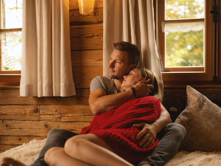 Paar liegt Arm in Arm auf Sofa vor Fenster in Holzhütte nahe dem Luxushotel Hüttenhof