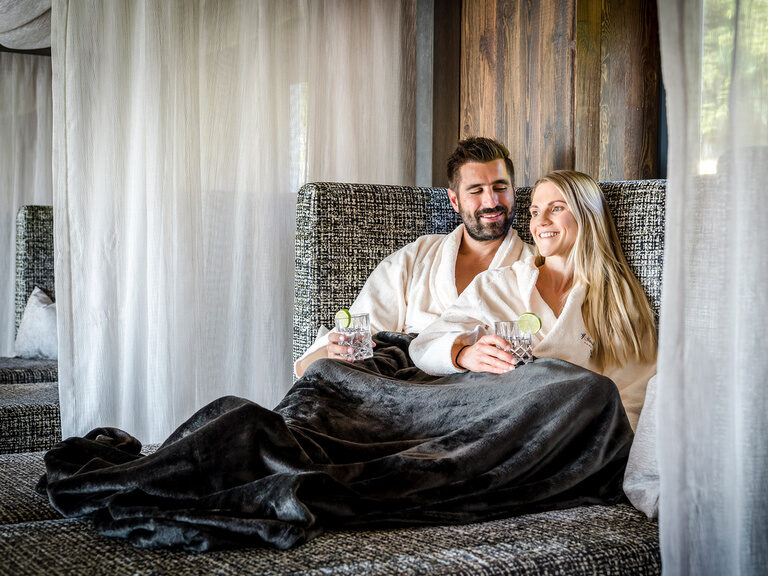 Paar liegt lachend mit Getränken im Bademantel auf Sofa-Liege mit Decke