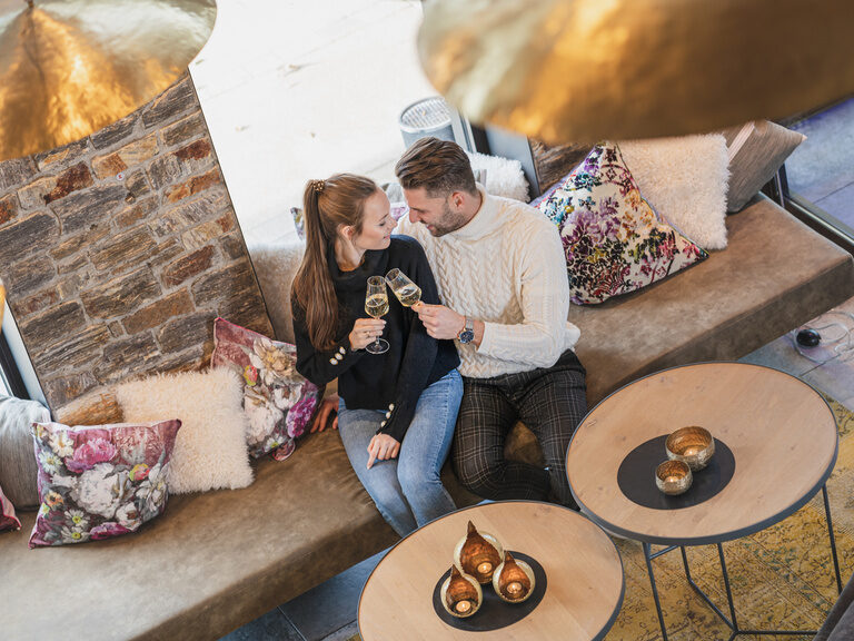 Paar sitzt lachend in Lounge-Bereich und stößt mit zwei Sektgläsern an