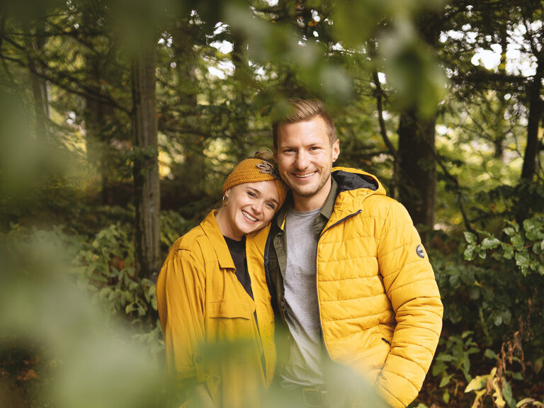 Paar steht lachend im Bayerischen Wald, Frau lehnt liebevoll Kopf an Schulter vom Mann
