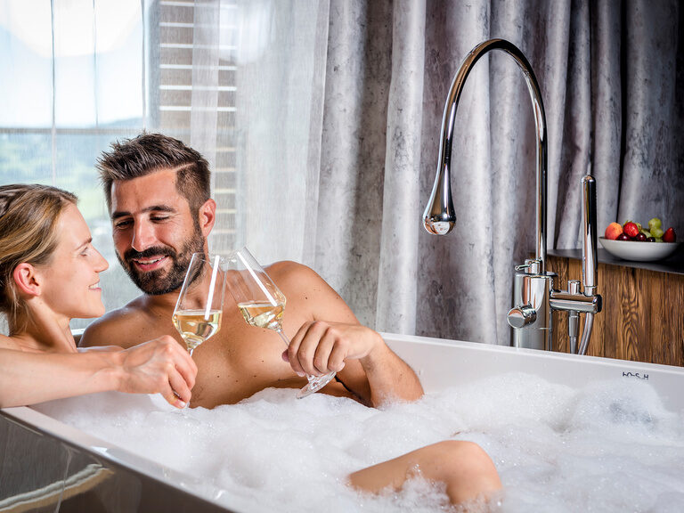 Paar lacht sich an und stößt in einer Badewanne mit zwei Sektgläsern an