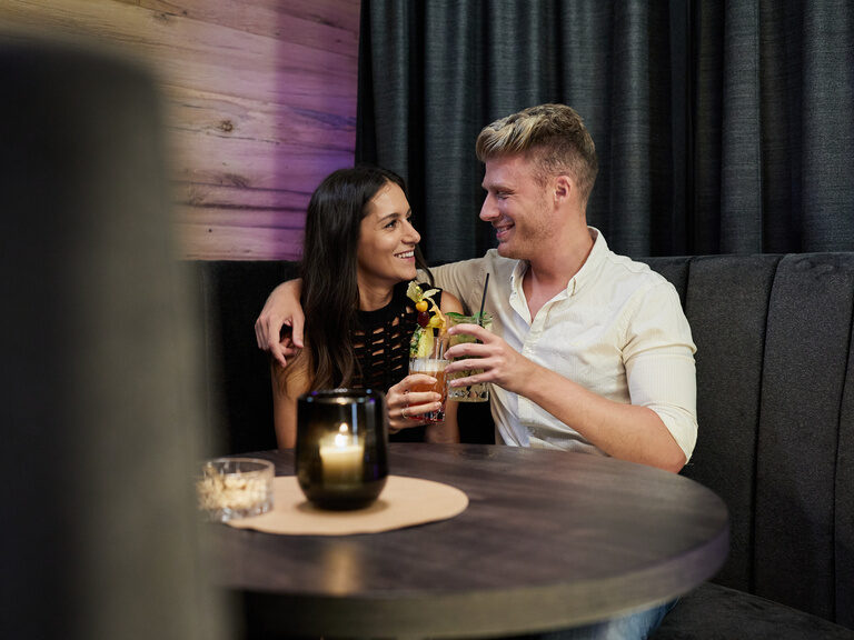 Paar stößt lachend und Arm in Arm am Tisch mit Cocktails an im Hotel Hüttenhof