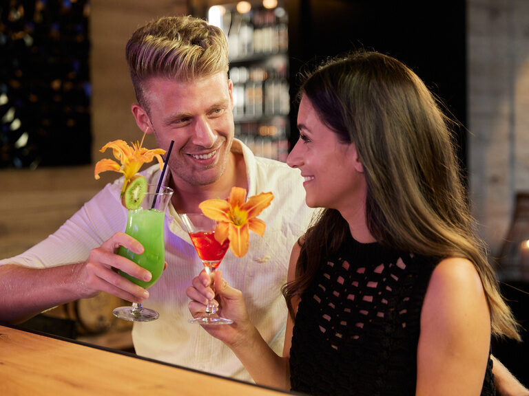Paar stoßt lachend mit zwei bunten, blumengeschmückten Cocktails in Bar vom Hüttenhof an