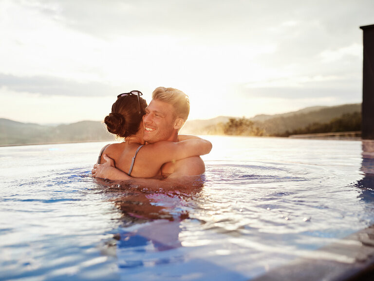 Pärchen umarmt sich bei Sonnenuntergang im Außenpool mit Blick auf die Berge