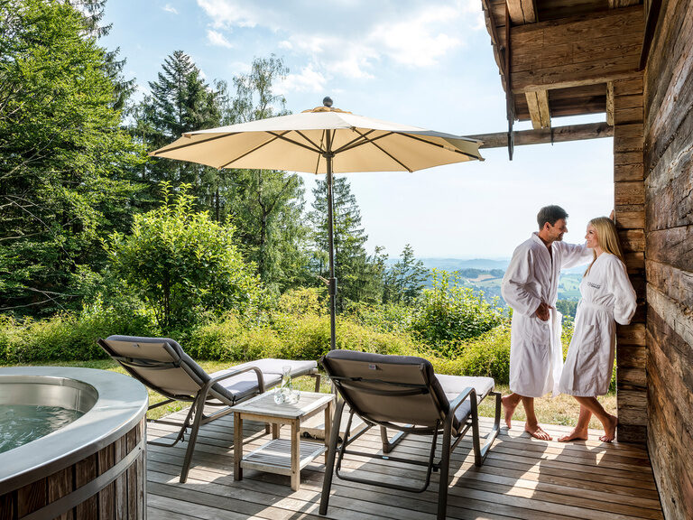 Pärchen steht auf der Terrasse des Chalets mit Whirlpool und gemütlichen Liegen, traumhafter Blick auf den bayerischen Wald