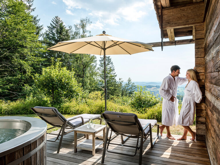 Bergdorf Chalet mit Whirlpool Hüttenhof Bayerischer Wald Pärchen steht auf der Terrasse des Chalets mit Whirlpool und gemütlichen Liegen, traumhafter Blick auf den bayerischen Wald